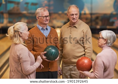 Waist up portrait of group of senior people holding bowling balls and chatting while enjoying active entertainment at bowling alley, copy space