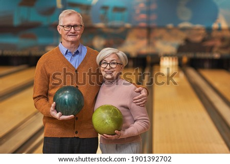 Waist up portrait of happy senior couple holding bowling balls and smiling at camera while enjoying active entertainment at bowling alley, copy space
