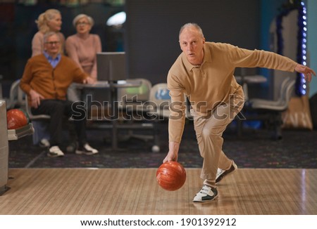 Full length portrait of active senior man playing bowling and throwing ball by lane with group of friends in background, copy space
