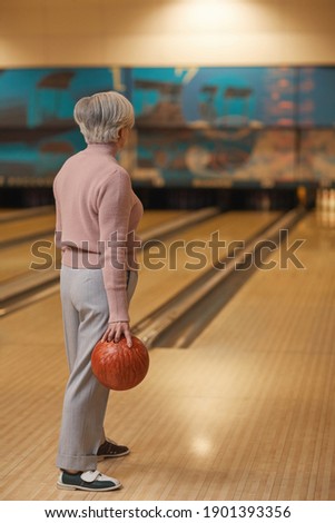 Vertical back view portrait of white haired senior woman playing bowling and looking away while standing by lane holding ball