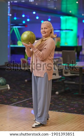 Vertical full length portrait of elegant senior woman holding bowling ball and smiling at camera while enjoying active entertainment at bowling alley