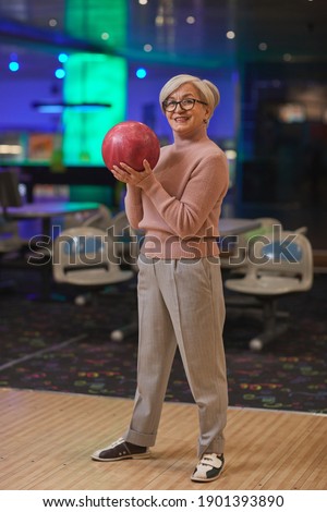 Vertical full length portrait of smiling senior woman holding bowling ball and looking at camera while enjoying active entertainment at bowling alley