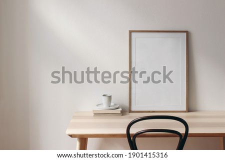 Breakfast still life scene. Cup of coffee, books and empty picture frame mockup on wooden desk, table. Elegant working space, home office concept. Scandinavian interior design. Room in sunlight.