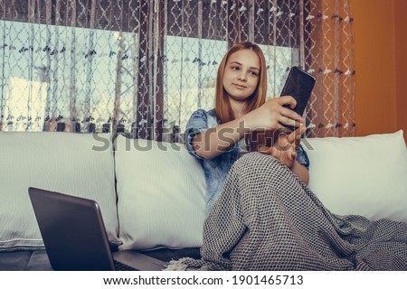 Beautiful teenage girl using laptop and taking selfie with her mobile phone while sitting covered with blanket in couch at home.
