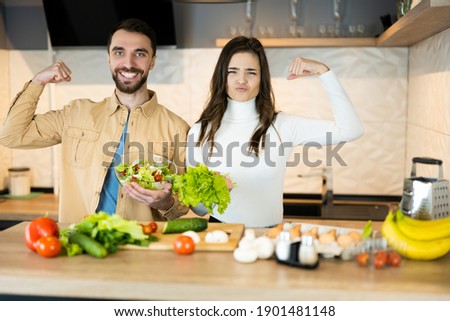 Cute vegetarian couple is showing how healthy and strong they are. Happy vegan people are cooking salad using only nutrition and healthy food Royalty-Free Stock Photo #1901481148