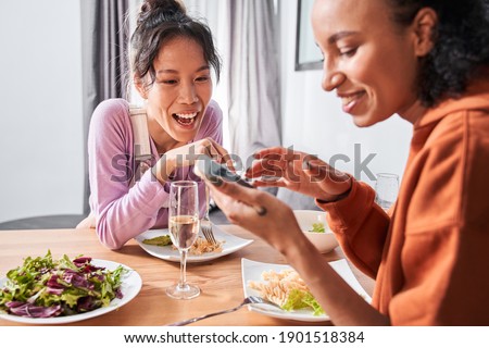 Portrait view of girls laughing while watching something at the smartphone during dinner in dining room. Glass of white wine standing at the table. Stock photo