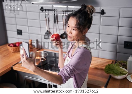 High angle view of a woman making selfie while eating apple. Beautiful smiling involved multiracial woman posing at the smartphone camera while standing at the kitchen. Stock photo