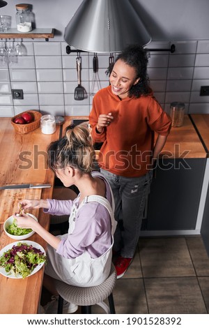 High angle view of the multiracial woman chatting with her bestie while she preparing salad at the table. Girl cooking together at the kitchen. Happy weekend of best friends concept. Stock photo