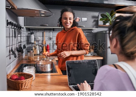 Back view of the woman sitting at the laptop and chatting with her multiracial best friend. Happy girls preparing pasta for breathtaking dinner. Coolinary concept. Stock photo