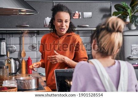 Happy girls preparing pasta for breathtaking dinner. Back view of the woman sitting at the laptop and chatting with her multiracial best friend. Coolinary concept. Stock photo
