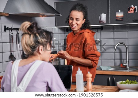 Happy curly woman telling something to her best friend while steering pasta at the pan. Girl spending time at the kitchen while preparing dinner. Coolinary concept. Stock photo