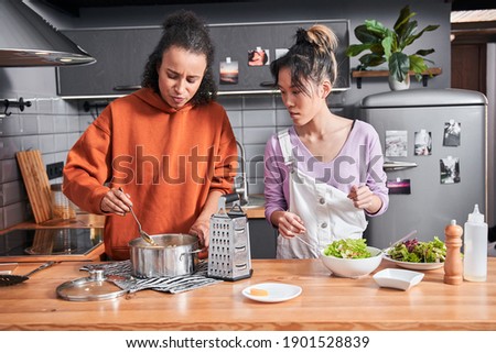 Serious attractive curly woman mixed pasta with cheese at the pan while preparing dinner for herself and her best friend. Her bestie looking at the dish and telling something. Stock photo