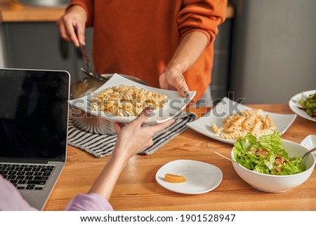 It for you. Cropped view of the multiracial woman giving plate with fresh pasta to her happy best friend sitting at the laptop. Two girls preparing to having dinner. Stock photo