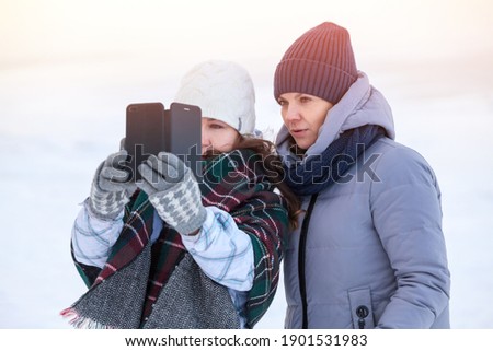 Two happy Caucasian women friends sharing a smart phone after photo self shooting in cold winter lake shore