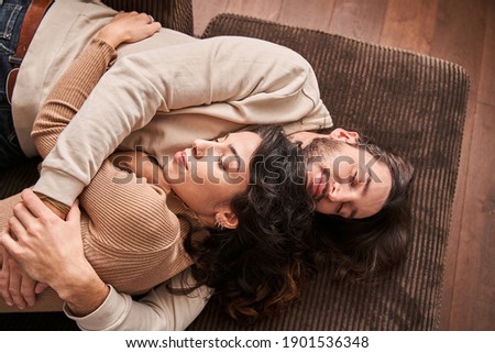 Love and tenderness. Image of optimistic smiling young loving couple laying on sofa indoors at home and hugging. Husband and wife enjoying of free time with each other. Stock photo