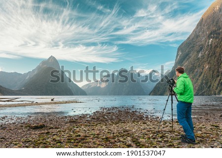 Tourist travel photographer taking pictures with professional camera on tripod on adventure travel vacation in mountain landscape. Milford Sound, Fiordland National Park, South Island, New Zealand.