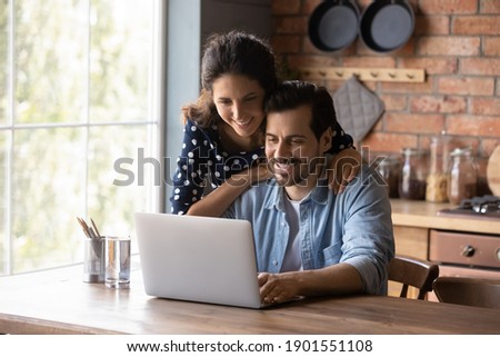 Smiling young Caucasian couple sit at table at home kitchen use laptop browsing wireless internet on gadget. Happy millennial man and woman look at computer screen shop online on device together.