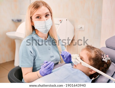 Close up dentist in medical face mask holding dental instruments while girl lying in dental chair with inhalation sedation at dental office. Concept of pediatric, sedation dentistry and dental care. Royalty-Free Stock Photo #1901643940