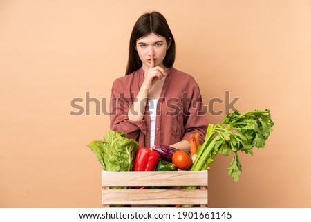 Young farmer girl with freshly picked vegetables in a box showing a sign of silence gesture putting finger in mouth