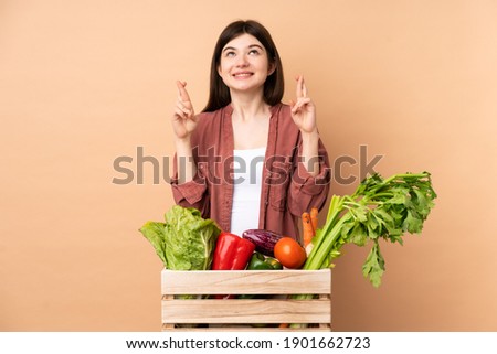 Young farmer girl with freshly picked vegetables in a box with fingers crossing and wishing the best