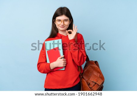Young Ukrainian teenager student girl holding a salad over isolated blue background showing an ok sign with fingers