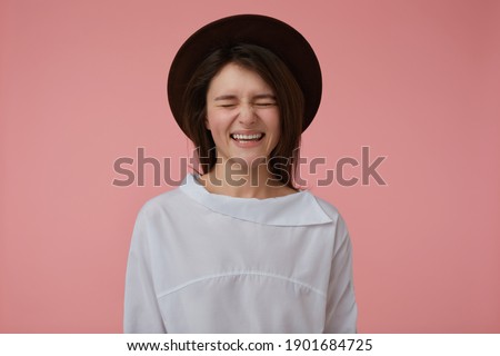 Teenage girl, happy looking woman with long brunette hair. Wearing white blouse and black hat. Laughing and keep eyes closed. Emotional concept. Stand isolated over pastel pink background