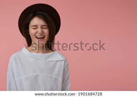 Portrait of attractive, adult girl with long brunette hair. Wearing white blouse and black hat. Laughing. Emotional concept. Stand isolated over yellow background, copy space at the right side