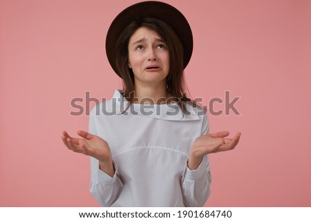 Upset looking woman, unhappy girl with long brunette hair. Wearing white blouse and black hat. Emotional concept. Stand isolated over pastel pink background and watching up at copy space