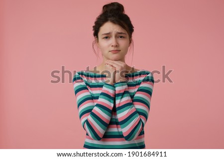 Portrait of asking, sad looking girl with brunette hair and bun. Wearing striped blouse and hold hands under her chin. Emotional concept. Watching at the camera isolated over pastel pink background