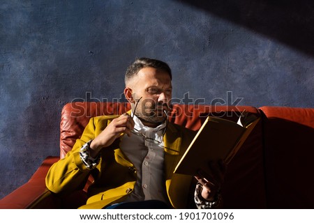 Stylish man with a beard in glasses reads a book sitting on an orange leather sofa on a background of dark blue wall. Wearing a white shirt and a yellow jacket. Training in self-isolation. Education.