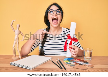 Brunette teenager girl artist holding painter diploma angry and mad screaming frustrated and furious, shouting with anger looking up. 
