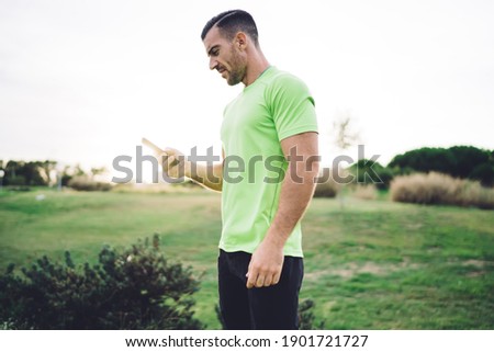 Backlit side view of Caucasian sportsman in active wear using cellphone application for browsing wireless internet, muscular male bodybuilder 30s checking received email from personal trainer