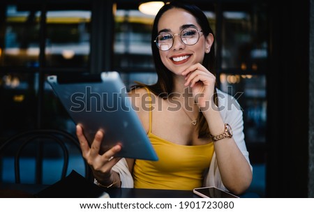 Portrait of cheerful Caucasian female blogger in eyewear smiling at camera during time for online networking via digital tablet, joyful hipster girl with modern touch pad posing in street cafe Royalty-Free Stock Photo #1901723080