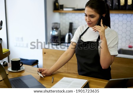 Skilled woman manage of cafeteria in apron making mobile phone call using number from visit card ordering supply, 20s smiling professional woman barista checking online booking via technology