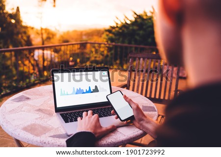 Cropped back view of business man using wireless internet connection on netbook and blank mobile for analyzing diagram, mockup smartphone gadget with copy space area for financial graph charts