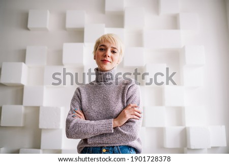 Low angle of glad female in casual clothes with short blond hair and folded hands looking at camera standing near wall with decorations