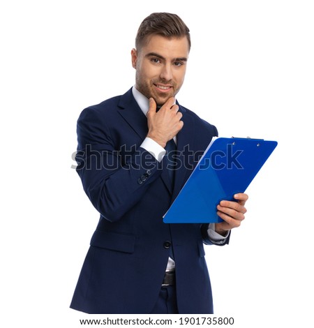 happy young model in elegant navy blue suit posing on white background, holding hand to chin, reading clipboard and smiling in studio