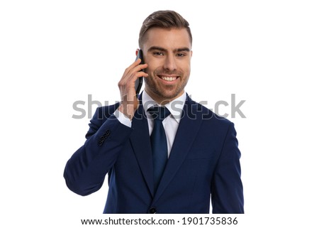 portrait of smiling young man in navy blue suit talking on the phone and standing isolated on white background in studio