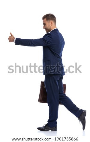 full body picture of unshaved businessman in navy blue suit making thumbs up sign, holding suitcase and walking isolated on white background in studio