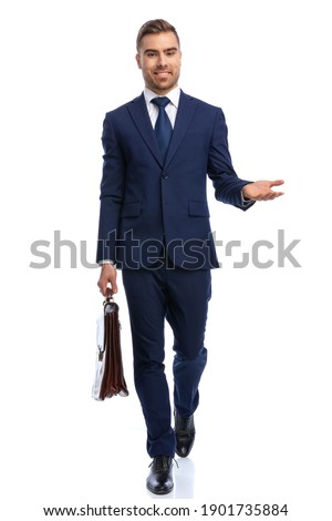 full body picture of attractive man in navy blue suit smiling, holding briefcase and presenting to side, walking isolated on white background in studio