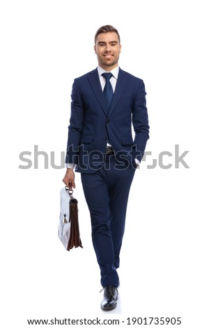 full body picture of smiling unshaved businessman in navy blue suit holding suitcase, walking with hand in pocket isolated on white background in studio