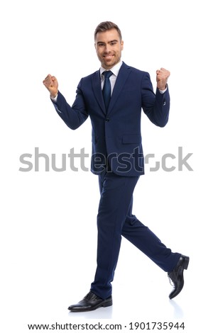 full body picture of happy young man in navy blue suit holding fists up, laughing and celebrating victory, walking isolated on white background in studio
