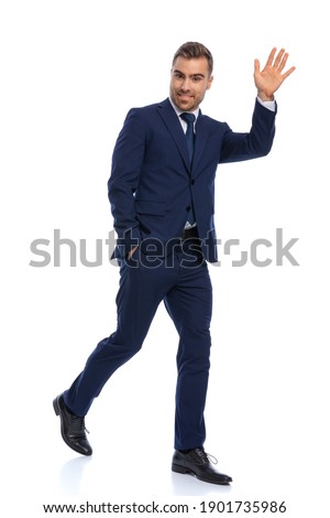 full body picture of happy businessman in navy blue suit holding hand up and waving, smiling and walking isolated on white background in studio