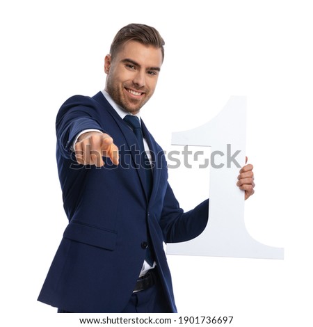 handsome unshaved elegant man in navy blue suit pointing finger and smiling, holding number one sign and posing isolated on white background in studio