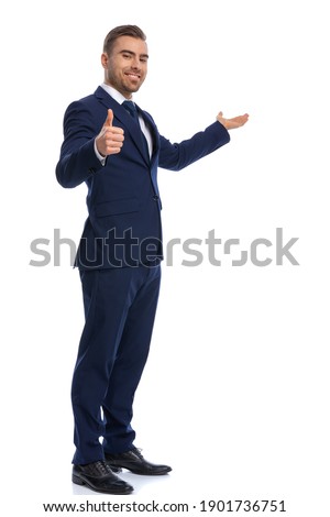 smiling unshaved guy in elegant suit making thumbs up sign and presenting to side, smiling and standing in a side view position isolated on white background, full body Royalty-Free Stock Photo #1901736751