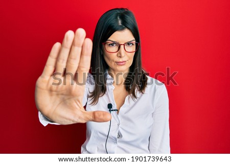 Young hispanic woman using lavalier microphone with open hand doing stop sign with serious and confident expression, defense gesture 