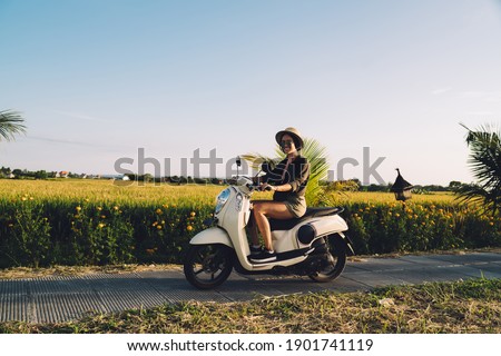 Portrait of cheerful female tourist sitting on rented vintage moped smiling at camera while getting to travel destination in Vietnam, funny woman in casual clothing ride on motorcy?le on path Royalty-Free Stock Photo #1901741119