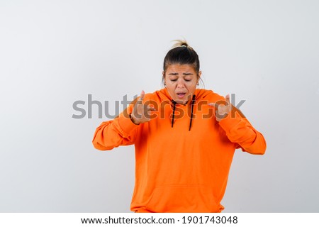  female pointing at herself in orange hoodie and looking alarmed. front view. 