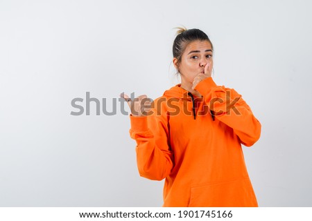  lady pointing aside with thumb, showing silence gesture in orange hoodie and looking sensible. front view. 