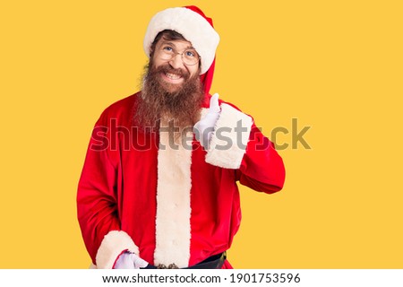 Handsome young red head man with long beard wearing santa claus costume doing happy thumbs up gesture with hand. approving expression looking at the camera showing success. 